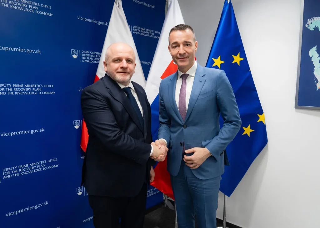 Zástupca Poľska a zástupca Slovenska si podávajú ruky počas oficiálneho stretnutia, v pozadí sú štátne a európske vlajky